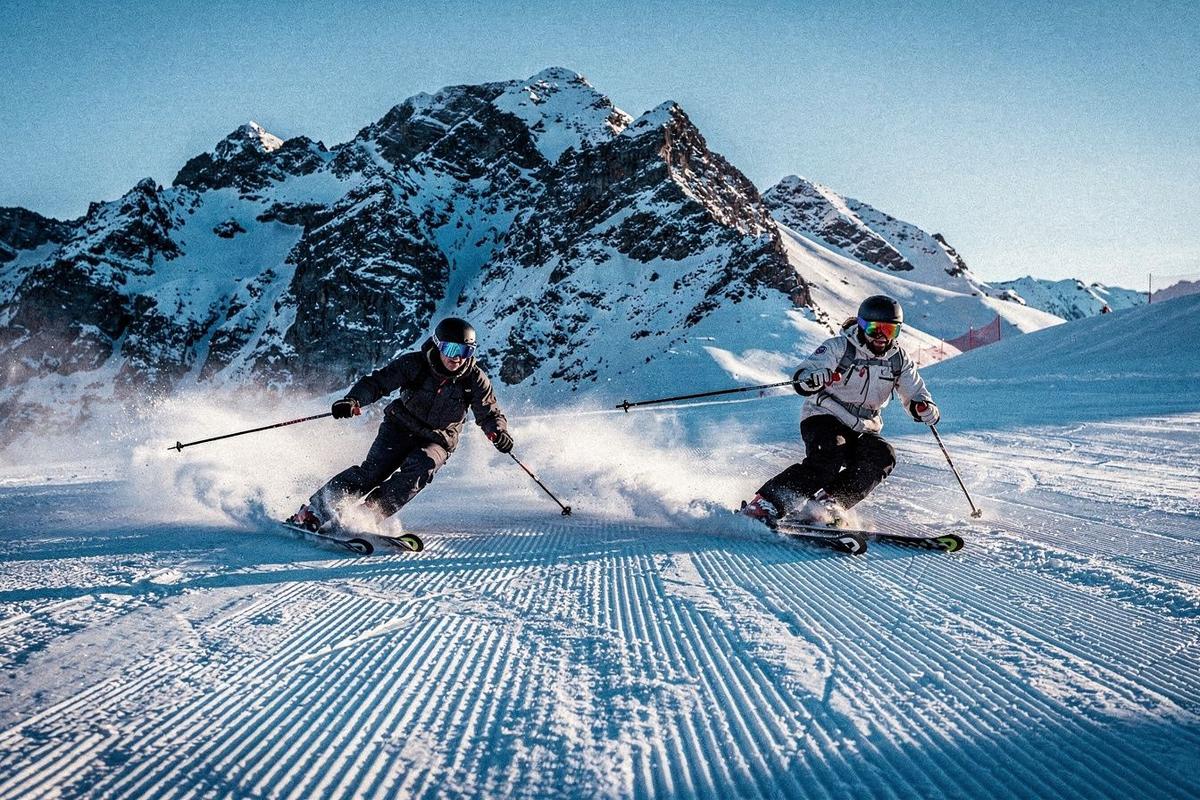 Two skiers carving a groomed piste below a snow-covered alpine ridge